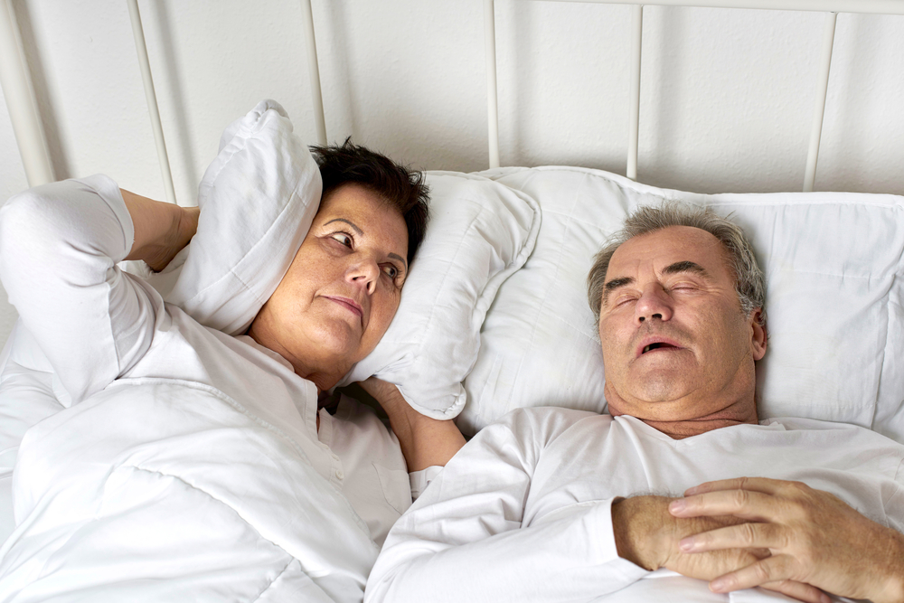 Eine Frau hält sich im Bett mit einem Kissen die Ohren zu, weil ihr Mann, der neben ihr liegt, laut schnarcht